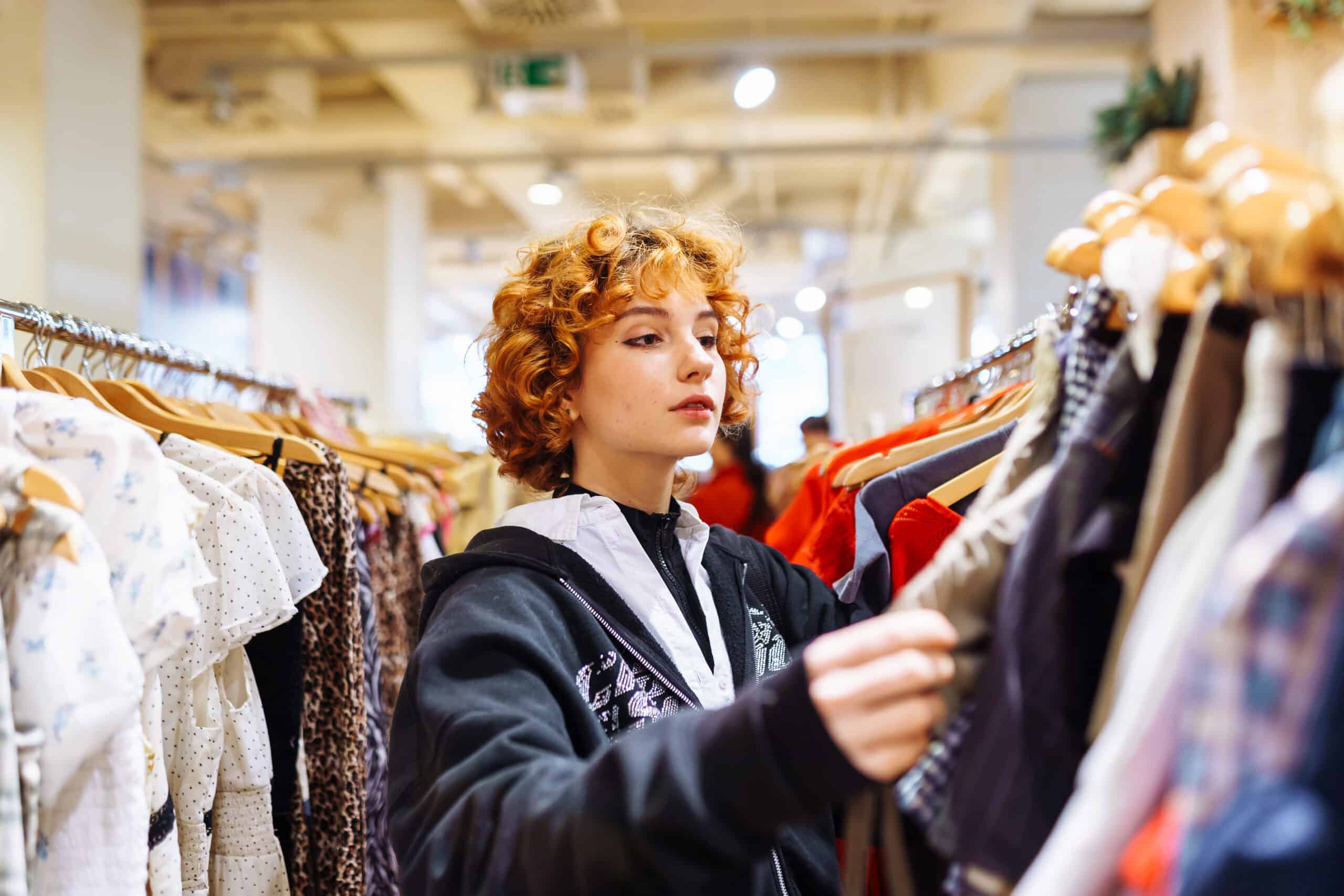 Woman with curly hair browsing clothes on a rack in a store aisle, focusing on garments nearby