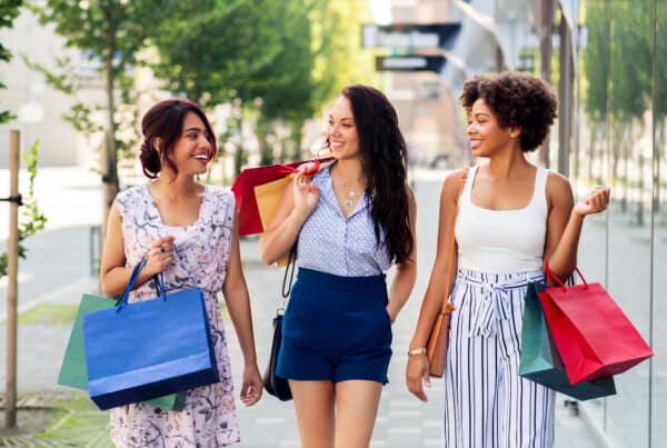 Three women carry colorful shopping bags and smile while walking on a sunny urban street.