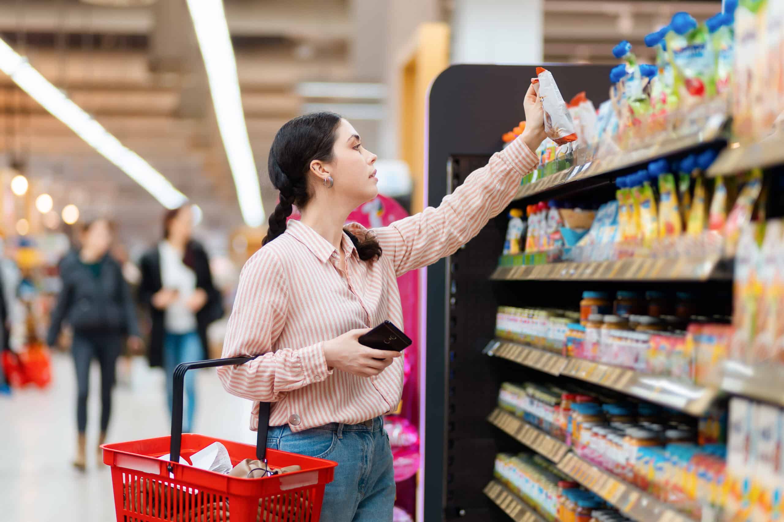 Woman in a striped shirt with a red shopping basket selects an item from a grocery store shelf.