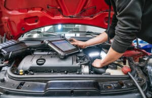 Mechanic uses a diagnostic tablet while the car hood is open, inspecting the engine bay under bright workshop lighting.