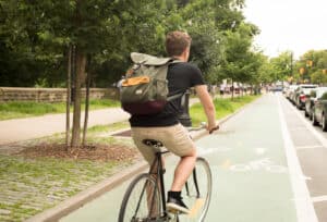 Back view of a cyclist riding in a green designated bike lane, wearing a large gray backpack and khaki shorts beside trees.