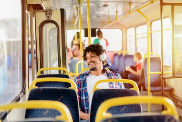Smiling man wearing white headphones sits on a bus with yellow handrails and other passengers in the background.