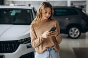 Woman in a beige sweater looking at her smartphone while standing beside a white SUV in a showroom or garage setting; soft focus background.
