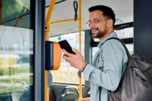 Man with backpack using a handheld device at a bus fare reader inside a vehicle.